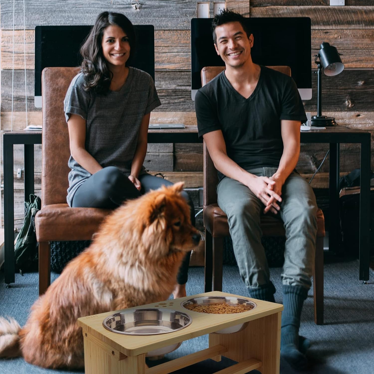 Two people sitting in a room with a dog and a pet feeding station.